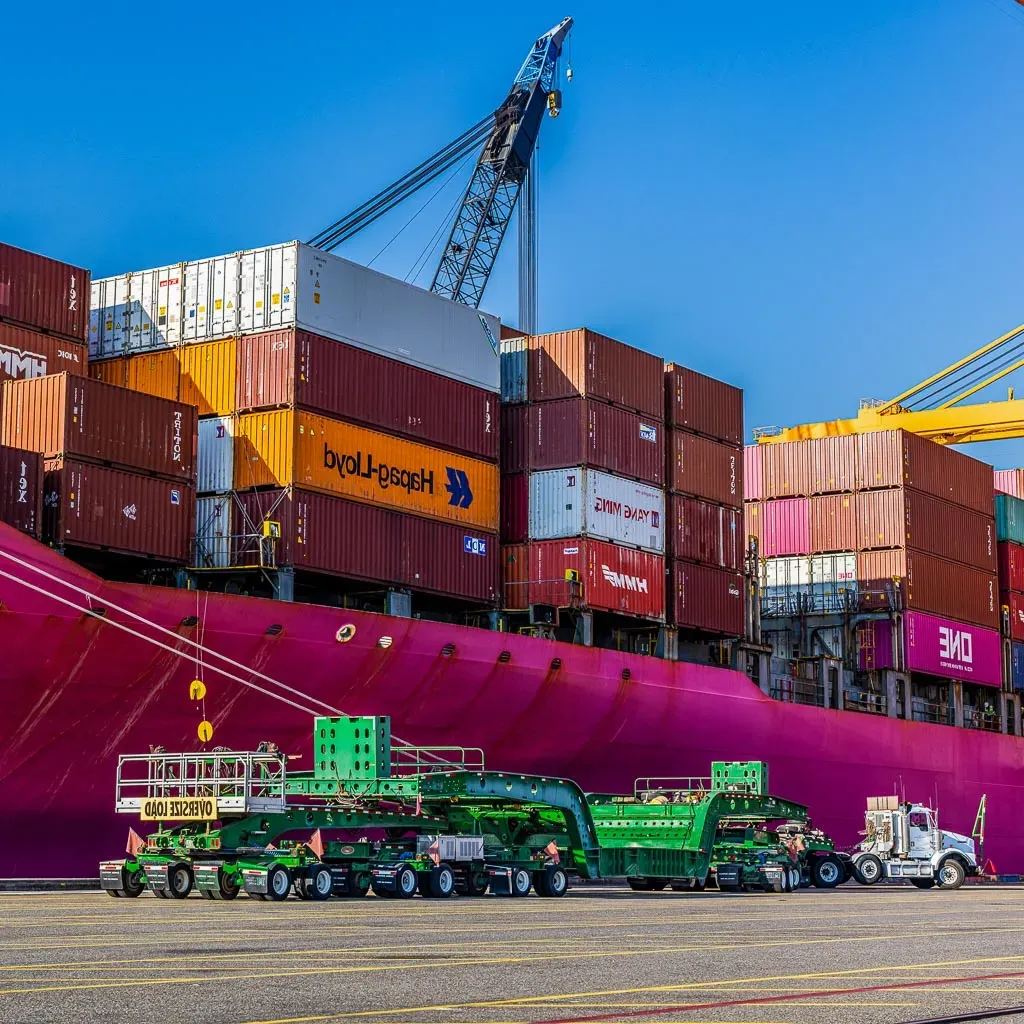 一辆欧米茄澳门威斯人平台首页双车道拖车停在一艘大型粉色集装箱船旁边.